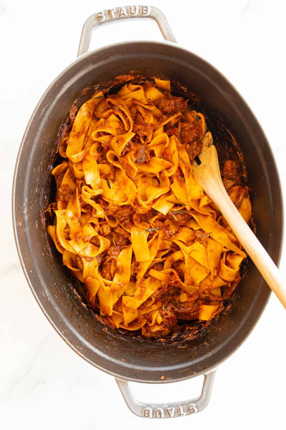 Beef ragu and Pappardelle pasta in a large cast iron pot, wooden spoon to the side.