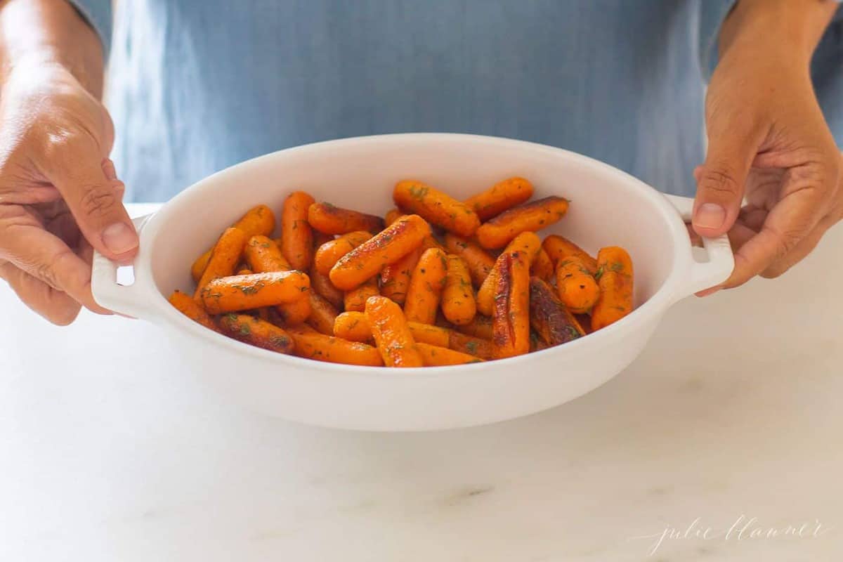 Woman in blue dress holding an oval baking dish filled with braised carrots. 