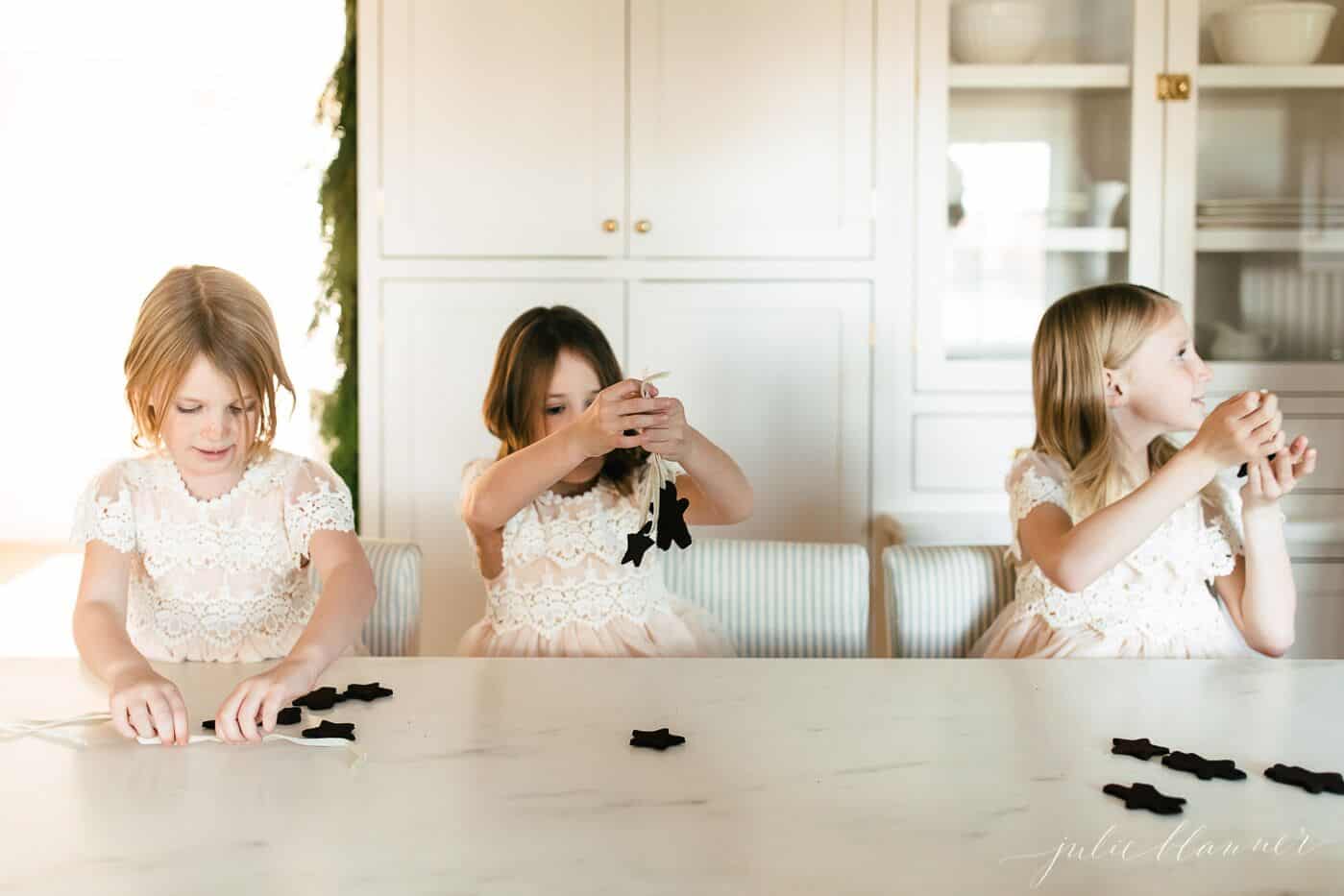 3 little girls threading ribbon through cinnamon applesauce ornaments.