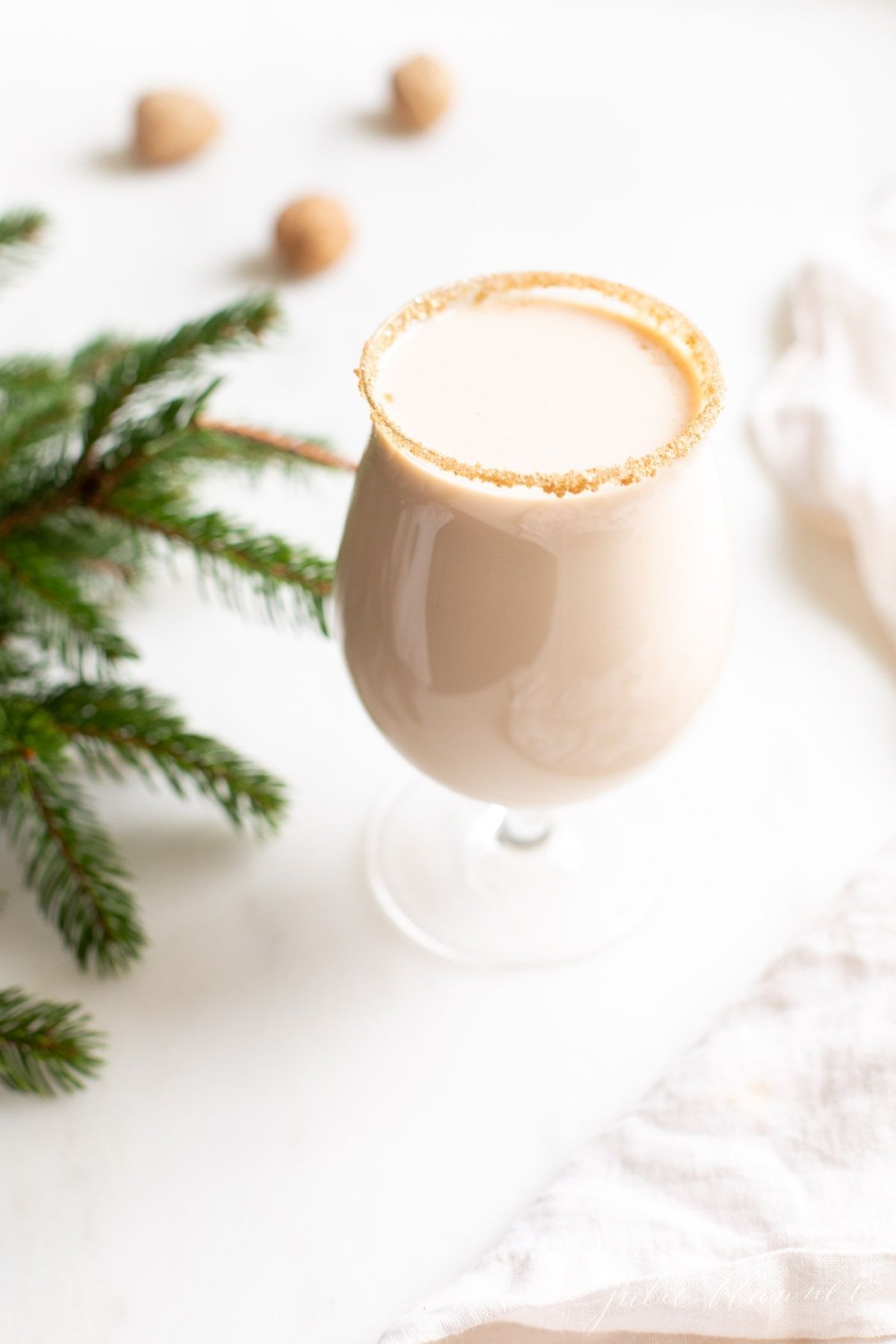 A clear glass with an oatmeal cookie cocktail, fresh winter greens on the edge of the image.