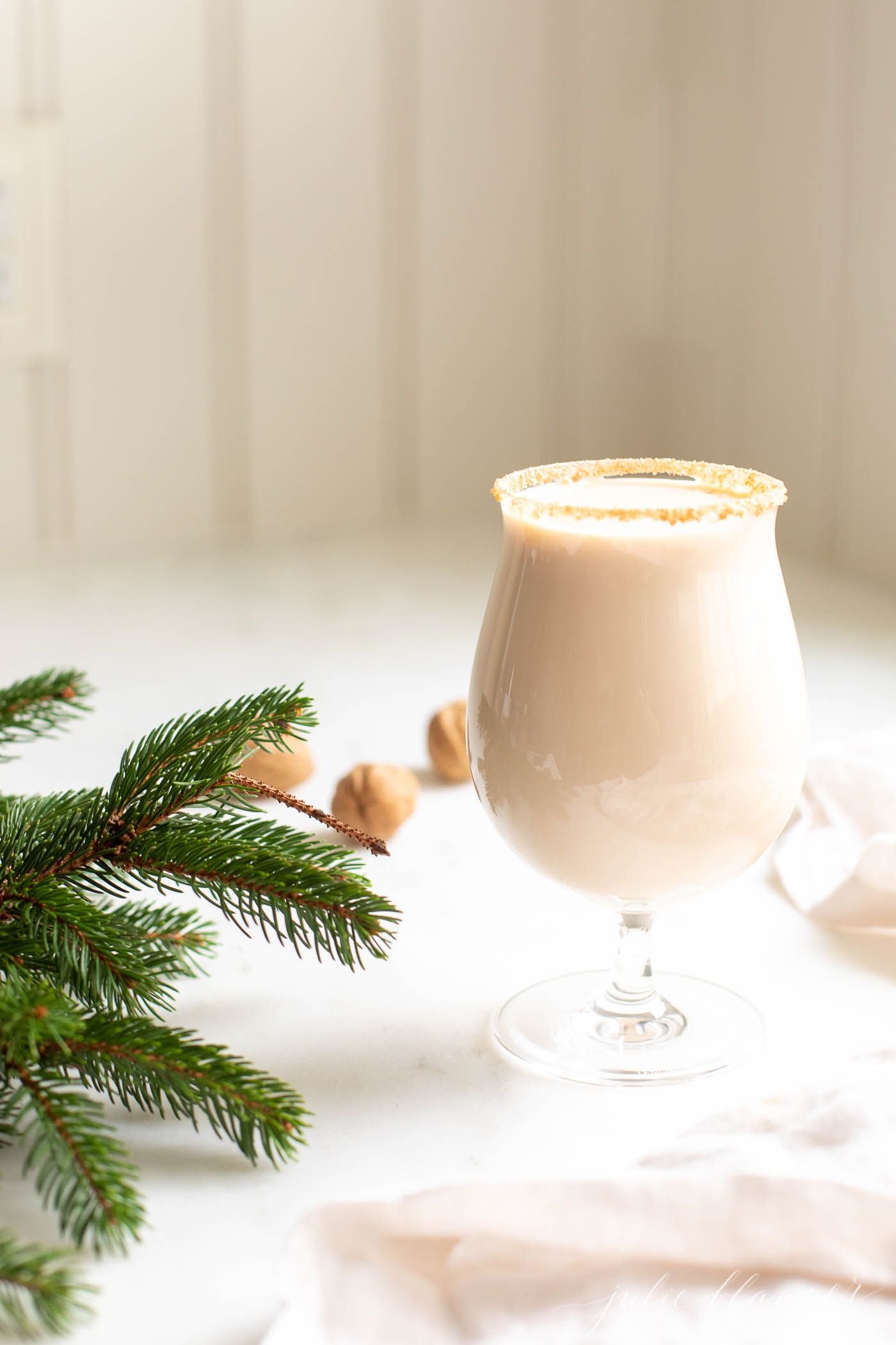 A clear glass with an oatmeal cookie cocktail, fresh winter greens on the edge of the image.