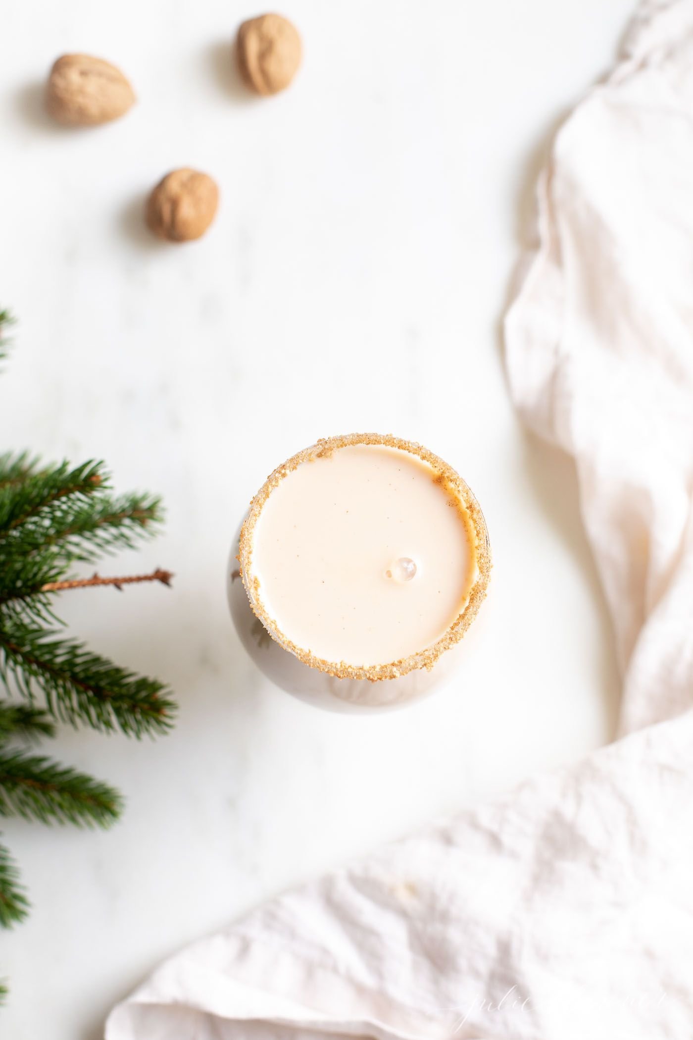 A clear glass with an oatmeal cookie cocktail, fresh winter greens on the edge of the image.