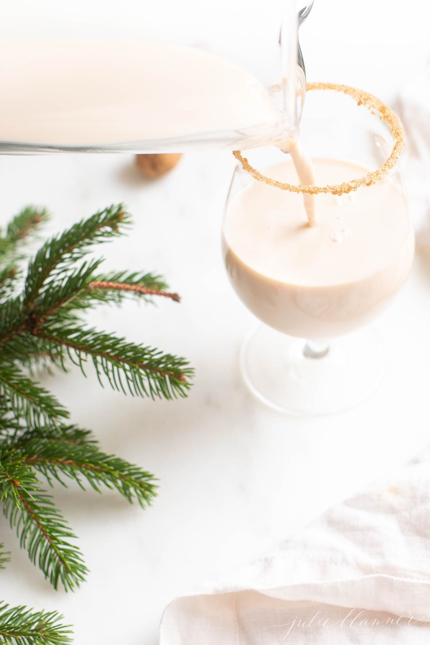 A clear glass with an oatmeal cookie cocktail, fresh winter greens on the edge of the image.