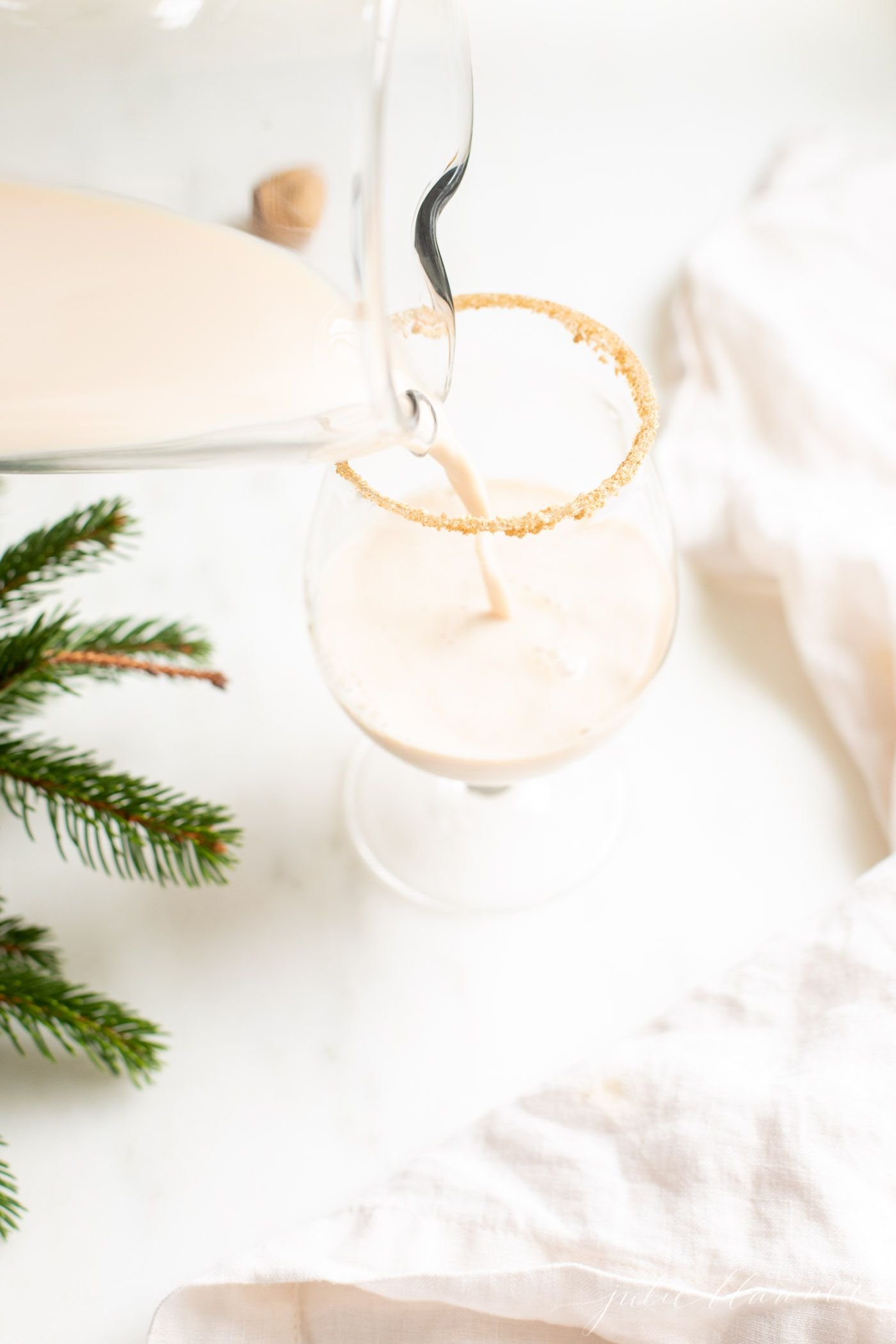 A clear glass with an oatmeal cookie cocktail, fresh winter greens on the edge of the image.