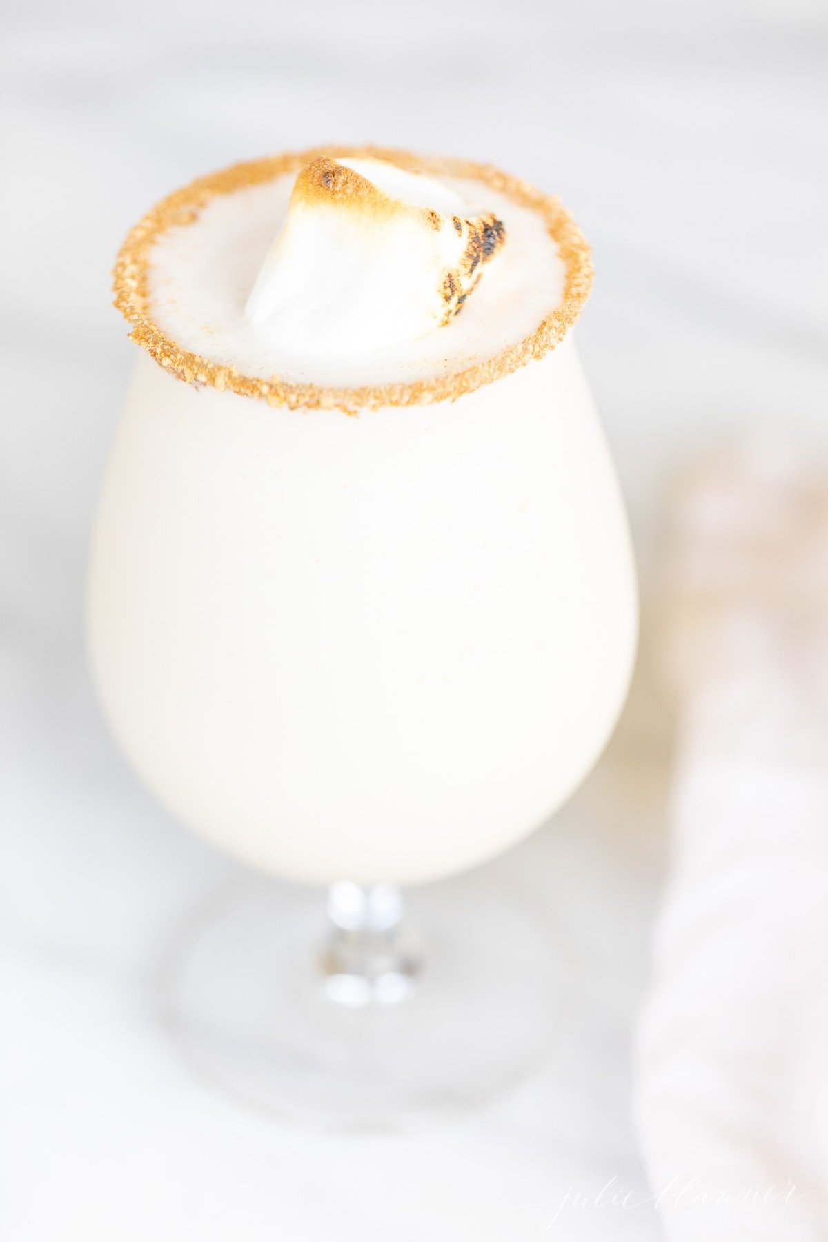 An ice cream and amaretto snowball cocktail in a stemmed glass, garnished with a toasted marshmallow, on a marble countertop.