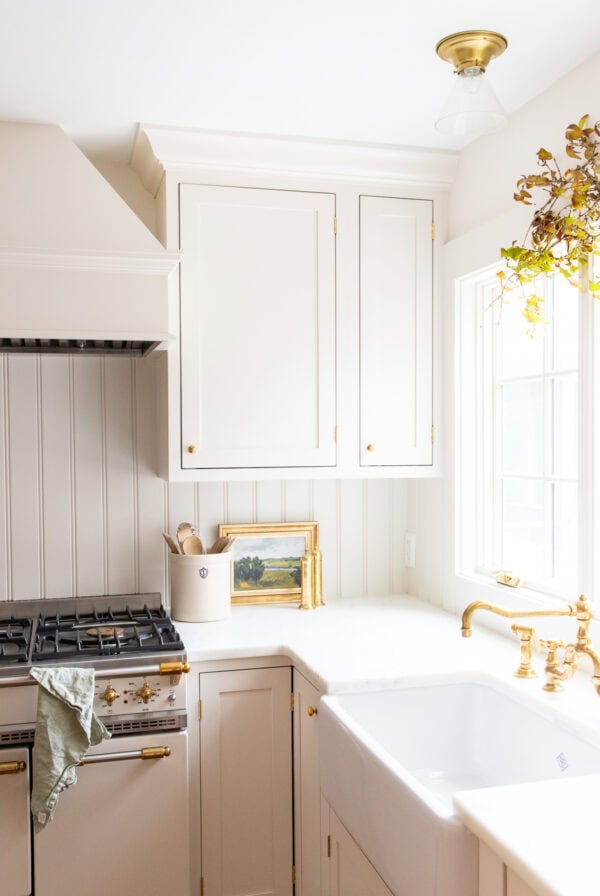 Bright kitchen with white cabinets, gold hardware, a farmhouse sink, and a gas stove—perfect for learning How to Host Thanksgiving. Natural light streams through the window; a small painting and utensils decorate the counter.