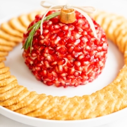 A pomegranate Christmas cheese ball shaped into an ornament, surrounded by crackers on a round white plate.