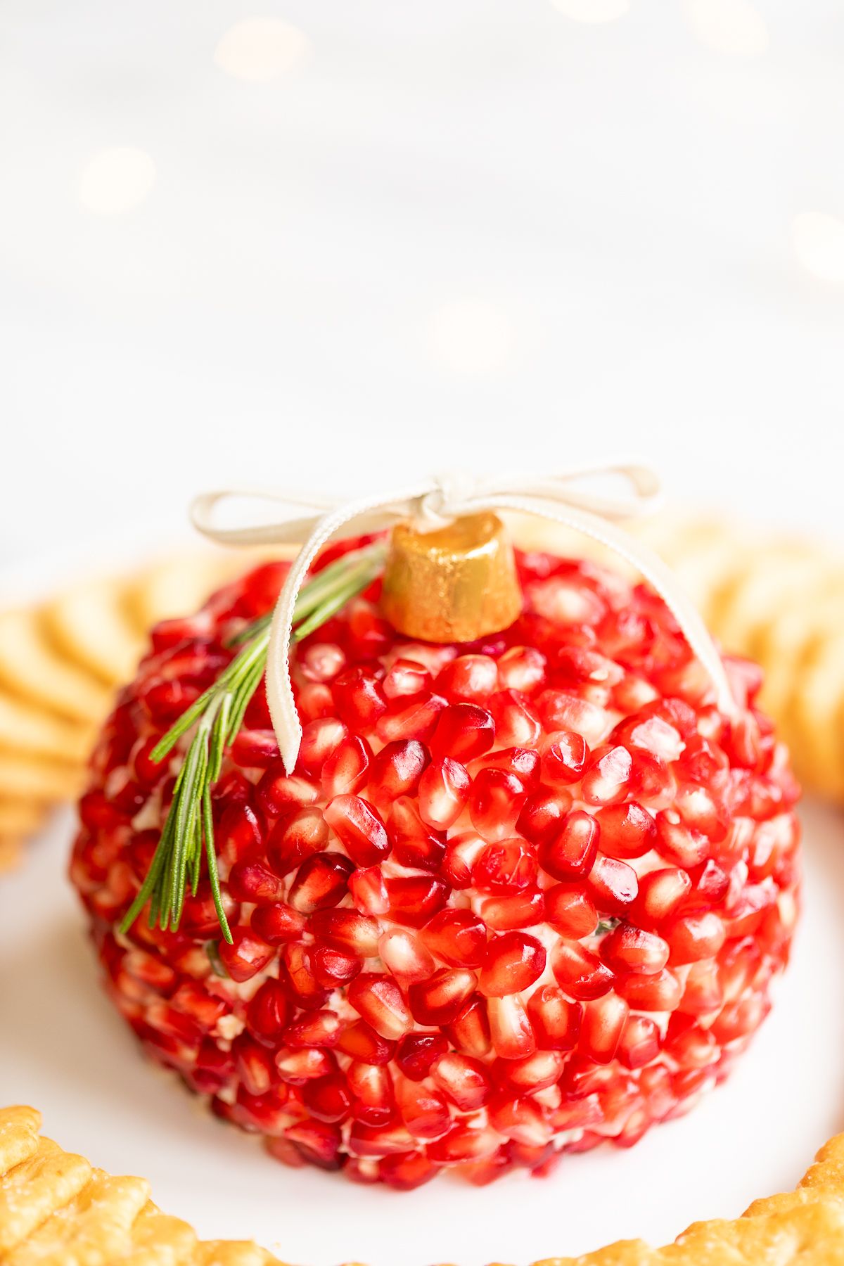 A pomegranate Christmas cheese ball shaped into an ornament, surrounded by crackers on a round white plate.