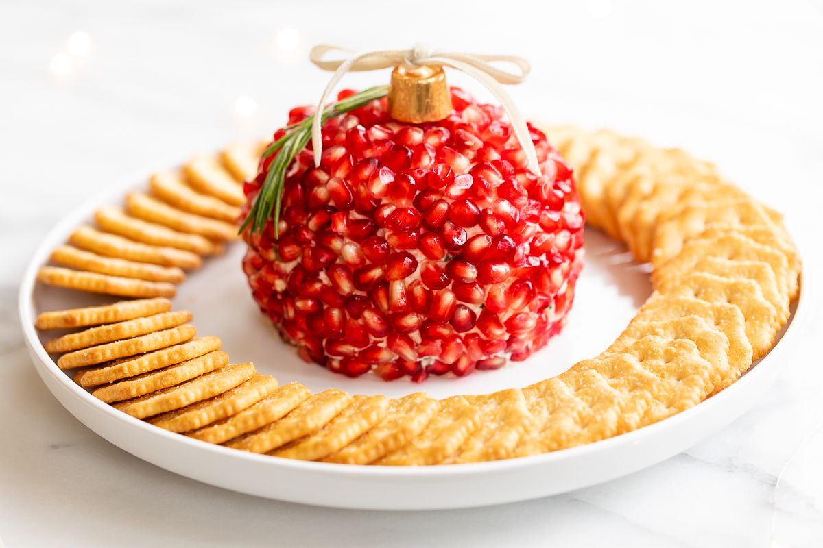 A pomegranate Christmas cheese ball shaped into an ornament, surrounded by crackers on a round white plate.