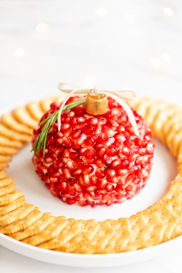 A pomegranate Christmas cheese ball shaped into an ornament, surrounded by crackers on a round white plate.