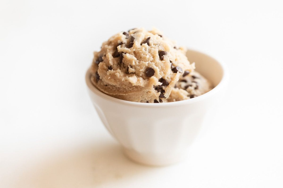 A small white bowl full of edible cookie dough with chocolate chips, on a marble surface