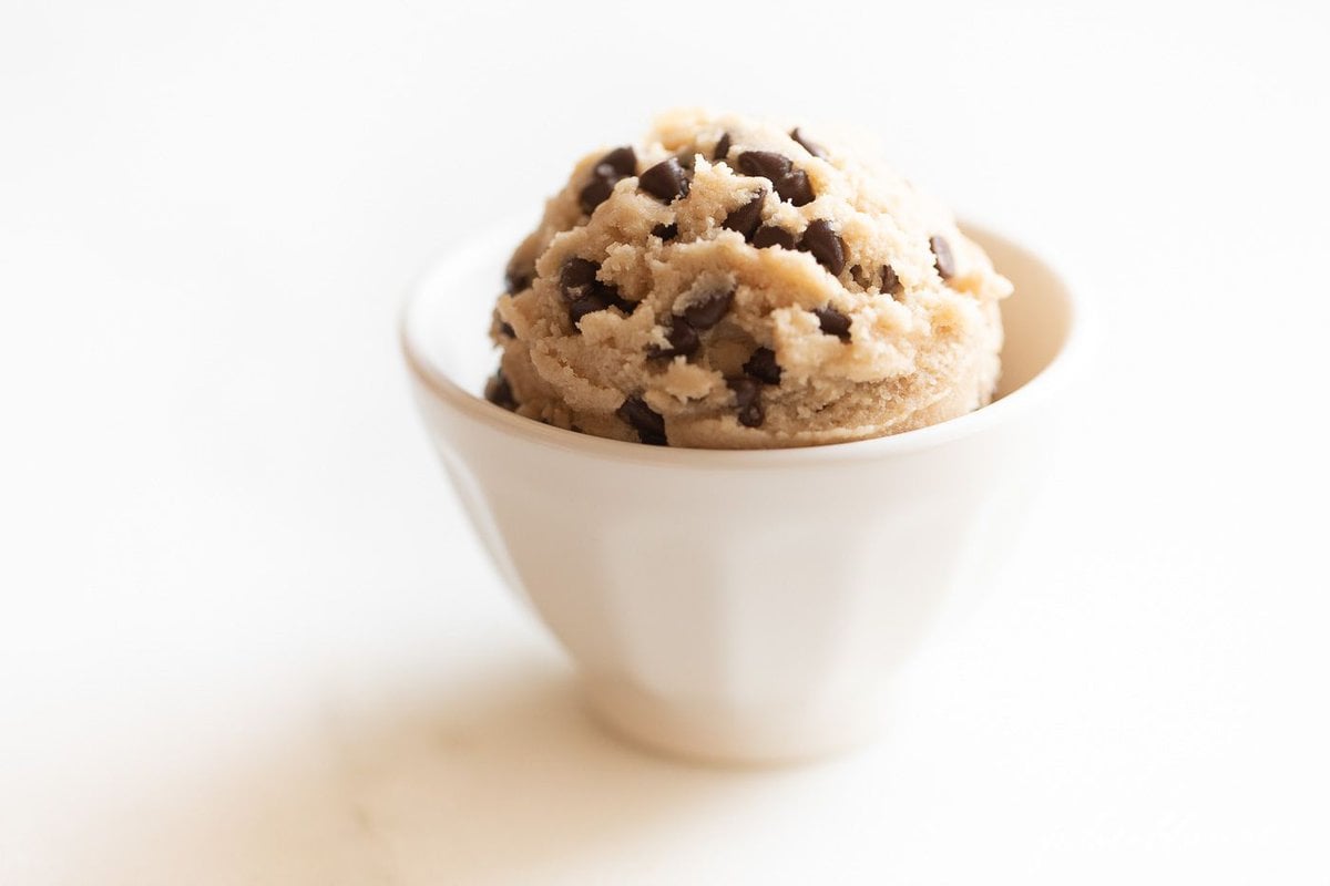 A small white bowl full of edible cookie dough with chocolate chips, on a marble surface