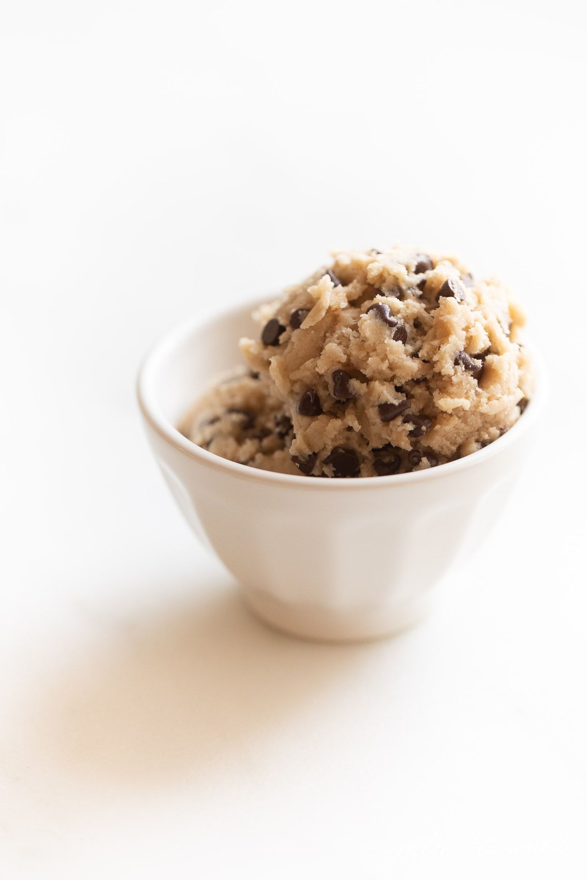 A small white bowl full of edible cookie dough with chocolate chips, on a marble surface