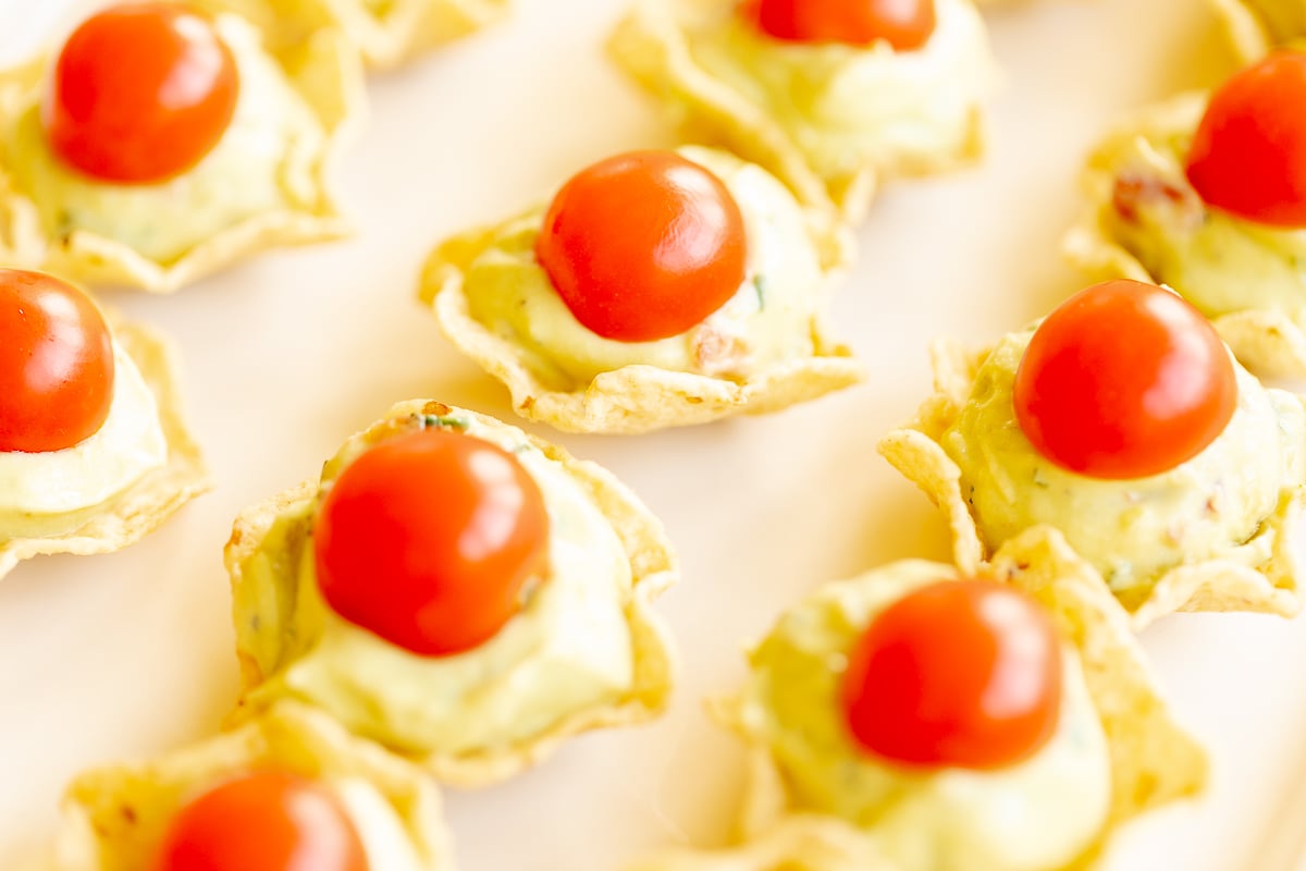 A white tray full of guacamole bites made from tortilla chips, topped with sliced cherry tomatoes.