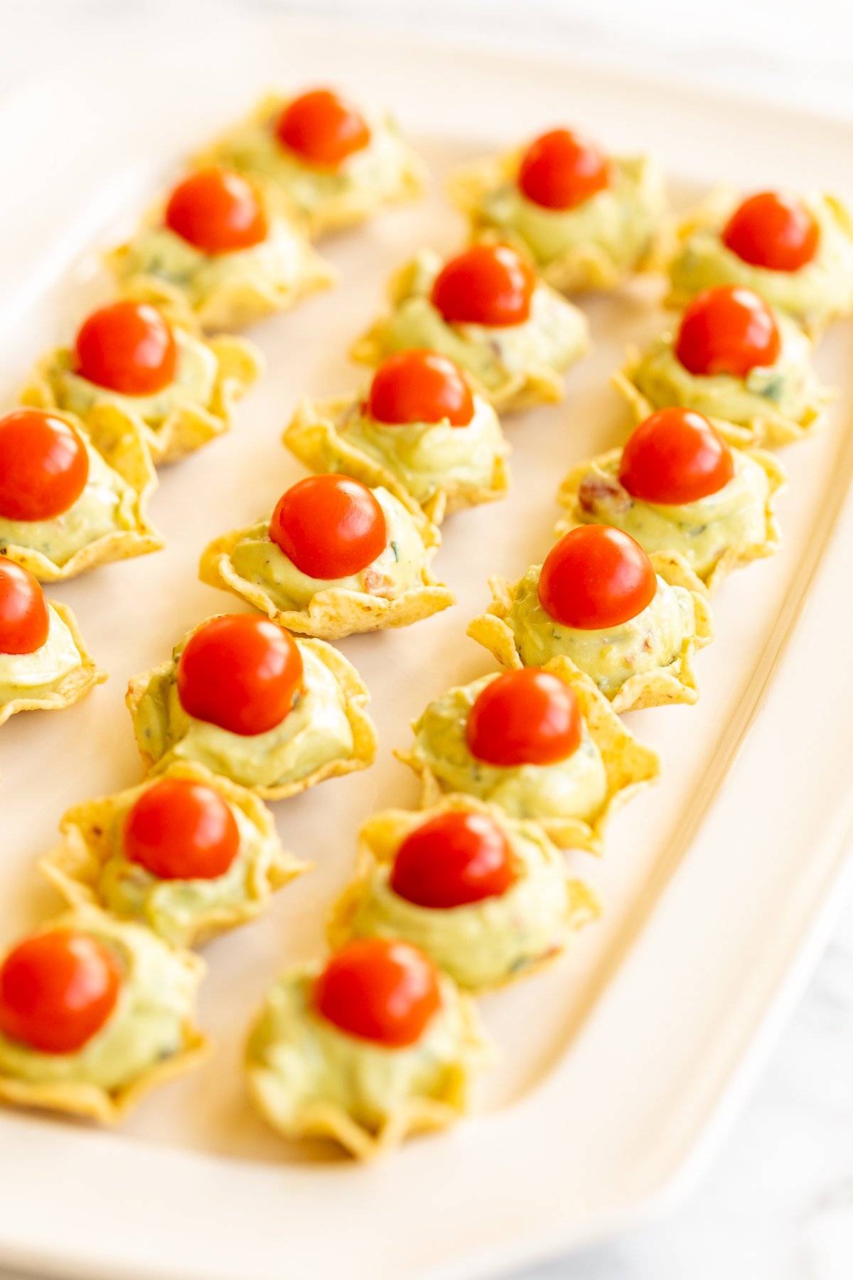 A white tray full of guacamole bites made from tortilla chips, topped with sliced cherry tomatoes.