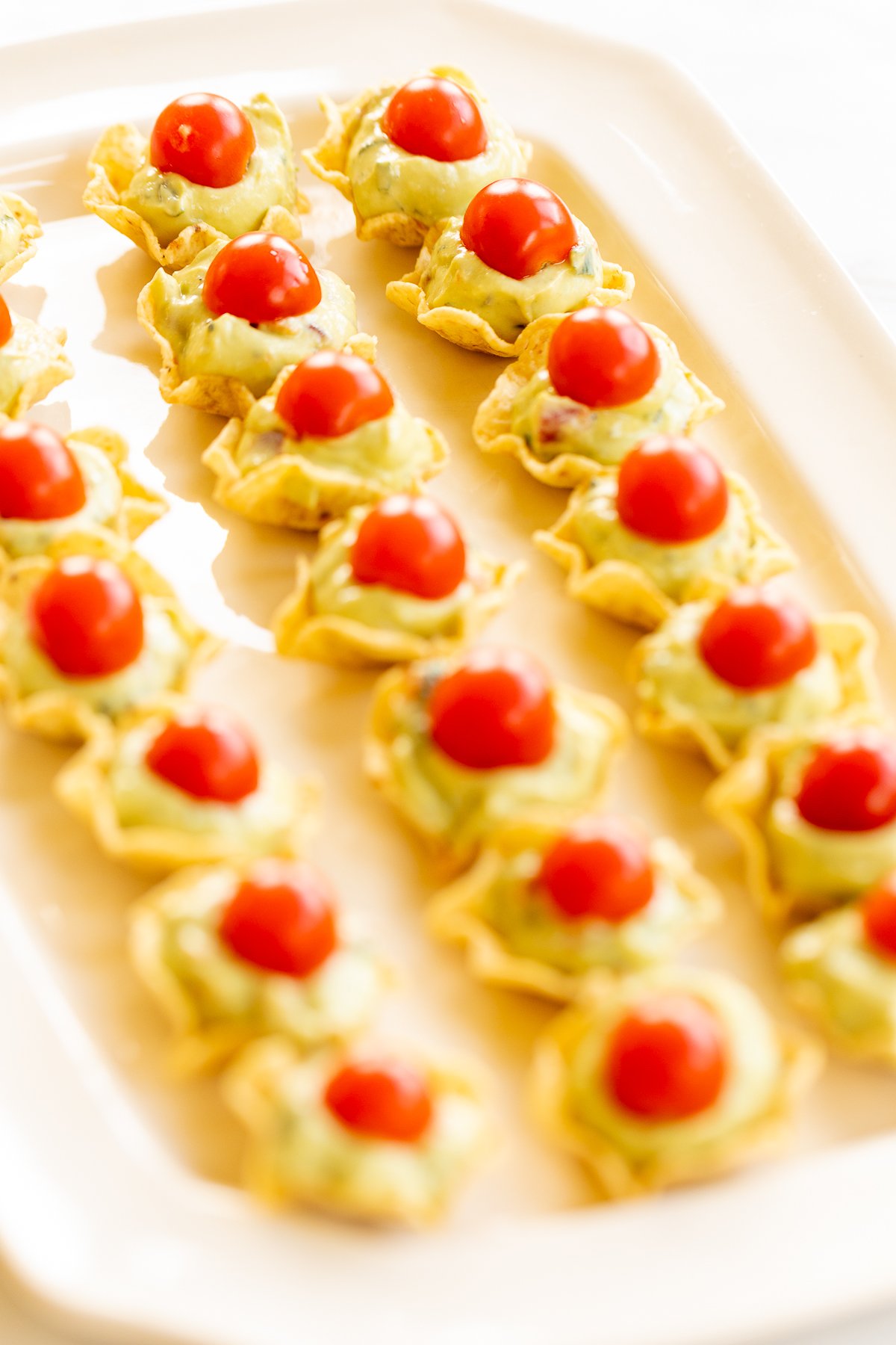 A white tray full of guacamole bites made from tortilla chips, topped with sliced cherry tomatoes.