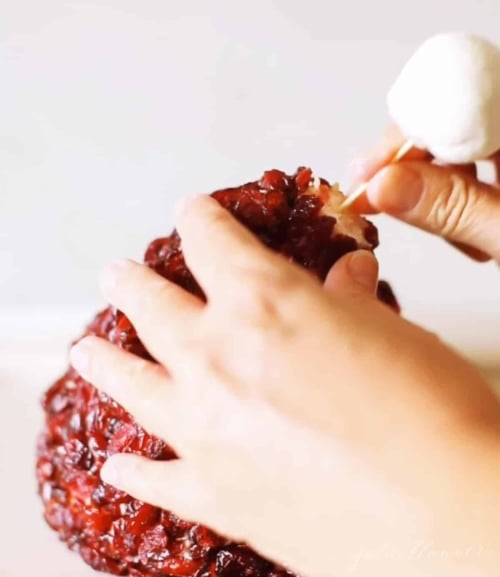 A hand placing a marshmallow on top of a cranberry covered cheese ball to make it look like a santa hat.