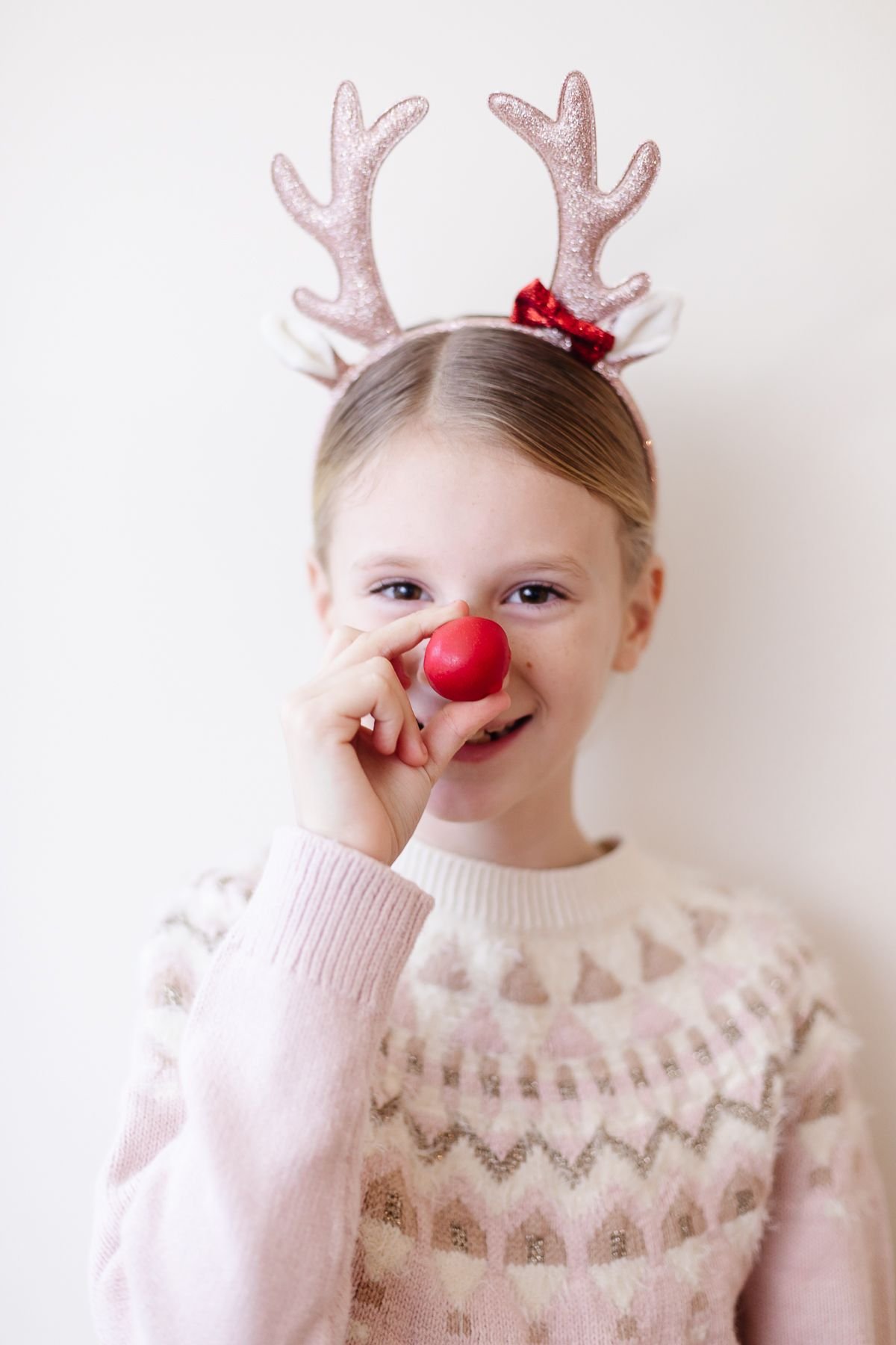 A little girl in reindeer antlers holding a red no bake Rudolph nose Christmas treat up to her nose.