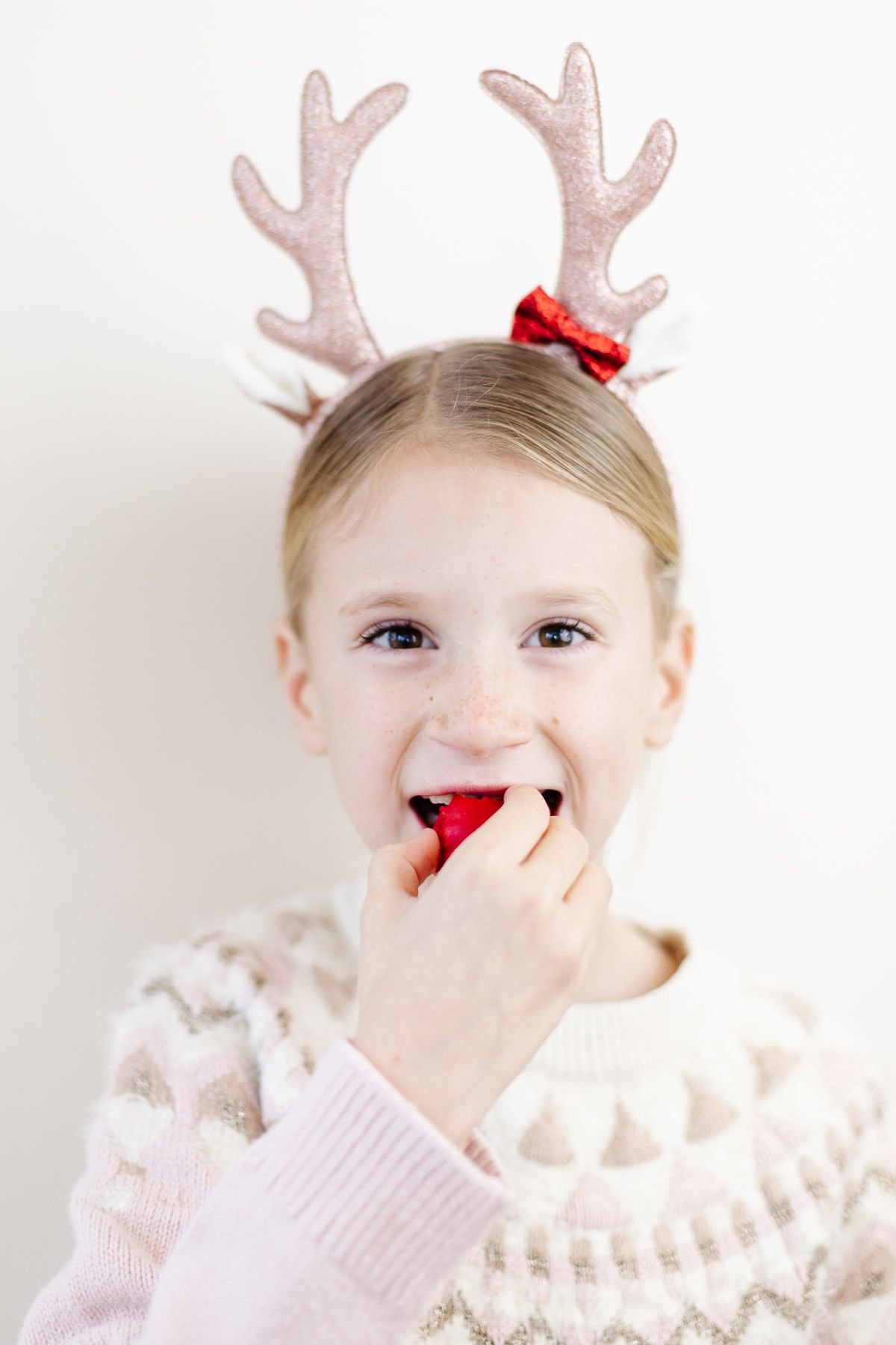 A little girl in reindeer antlers biting into a red no bake Rudolph nose Christmas treat.