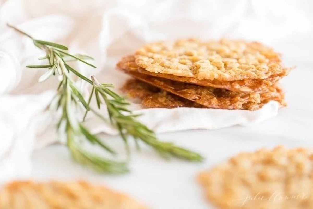 A stack of oatmeal lace cookies on a white surface, with a touch of greenery in the corner