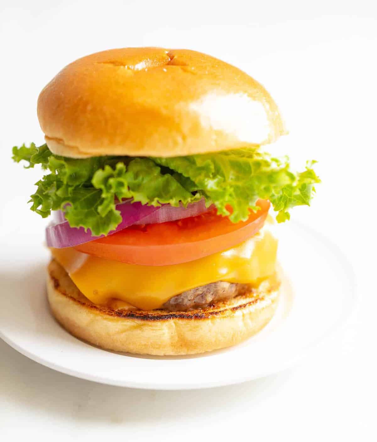 White background with a white plate featuring a classic cheeseburger, stacked with cheese, tomato, onion and lettuce.