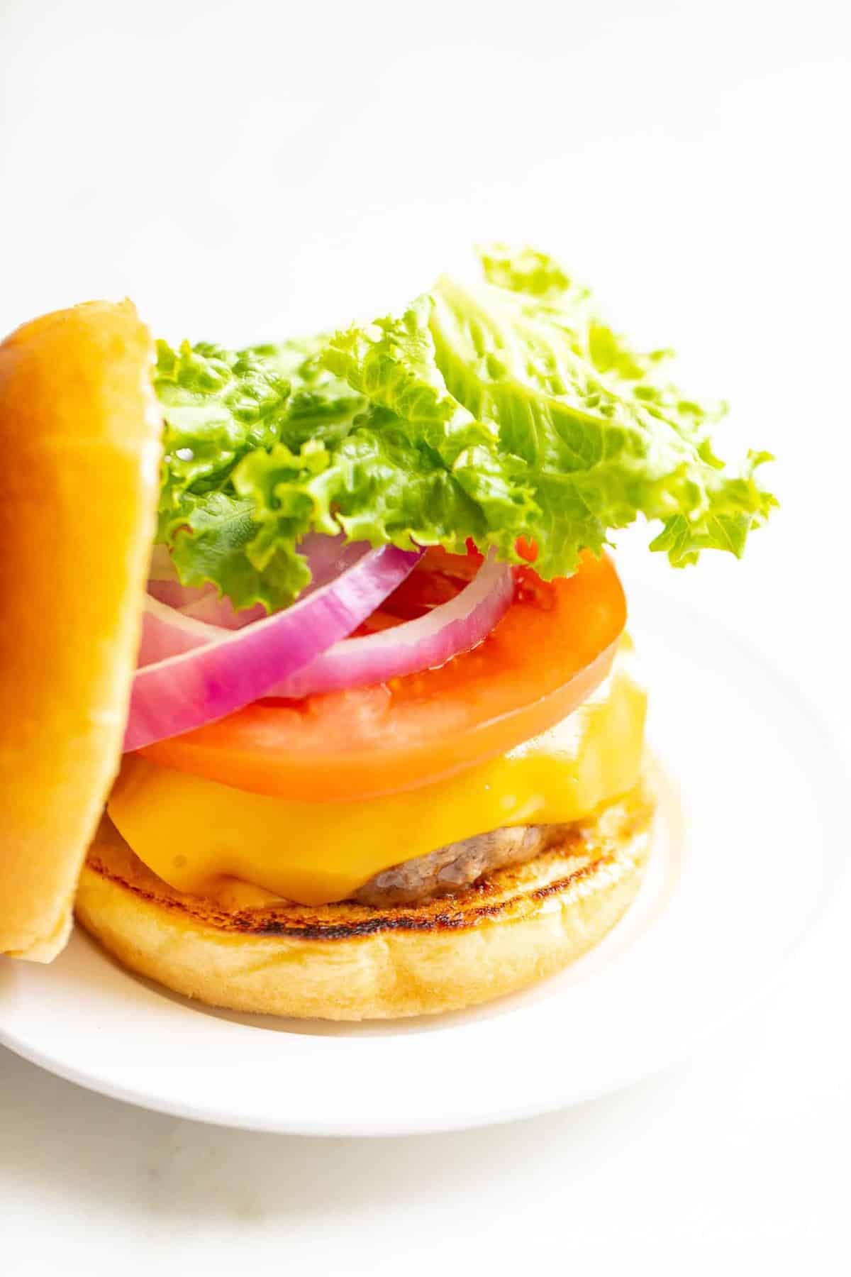 White background with a white plate featuring a classic cheeseburger, stacked with cheese, tomato, onion and lettuce, top bun to the side. 