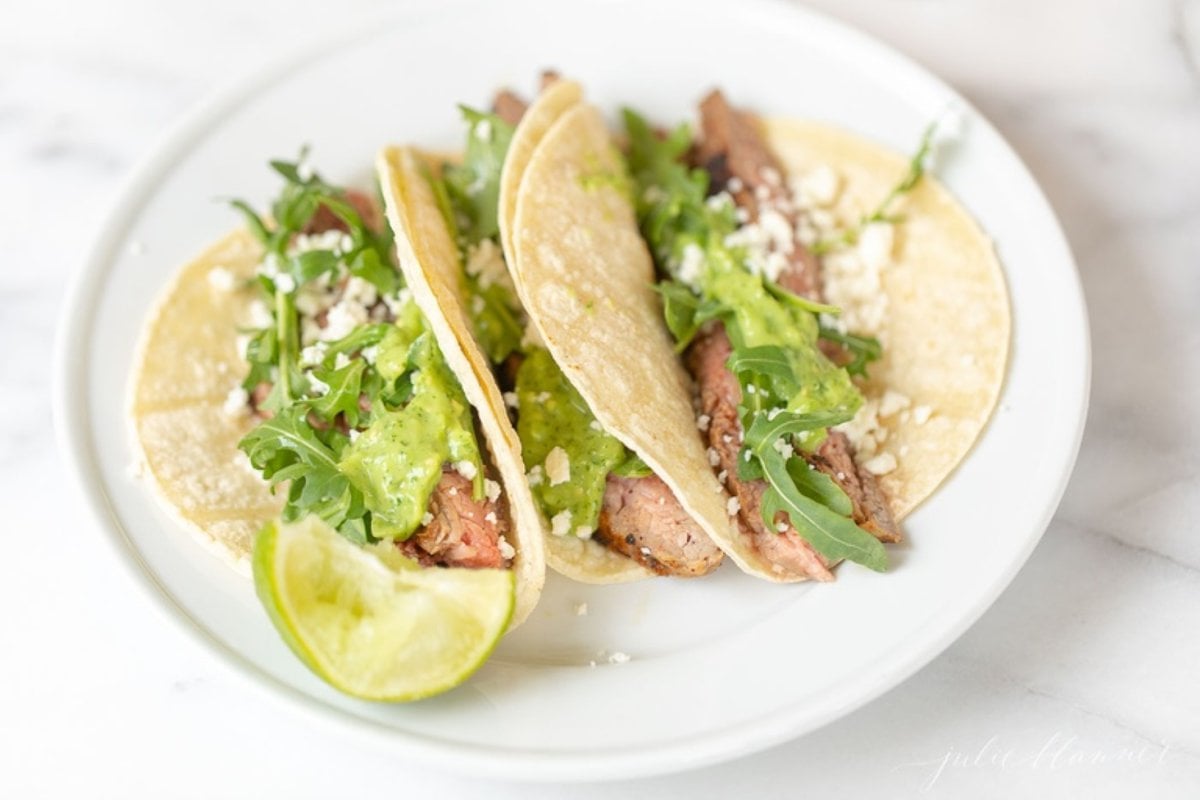 A white plate featuring three carne asada tacos topped with sauce, cheese and arugula. 