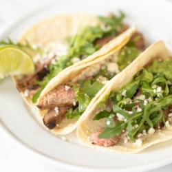 A white plate featuring three carne asada tacos topped with sauce, cheese and arugula.