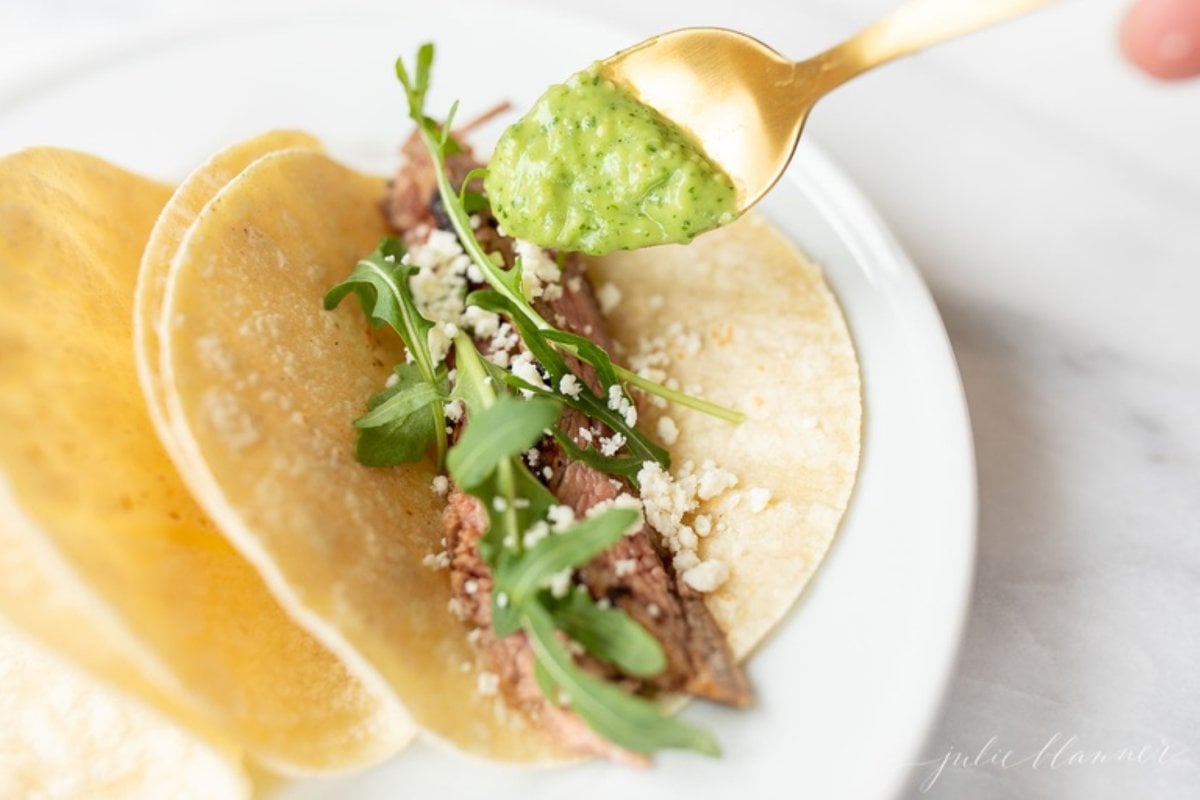 A hand holding a spoonful of green serrano sauce over a carne asada taco.