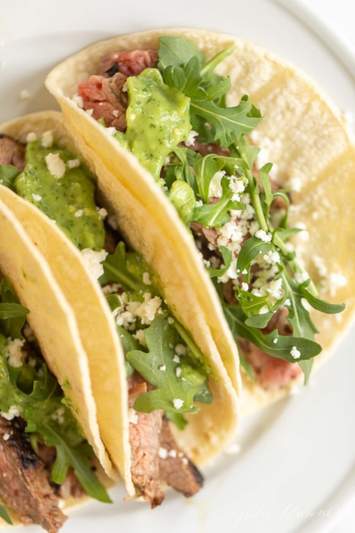 A white plate featuring three carne asada tacos topped with sauce, cheese and arugula.