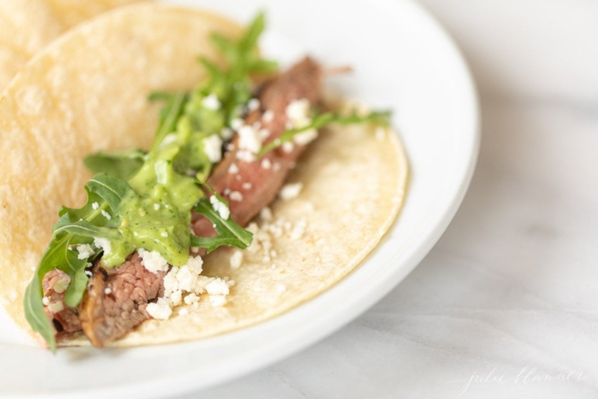 A white plate featuring three carne asada tacos topped with sauce, cheese and arugula.