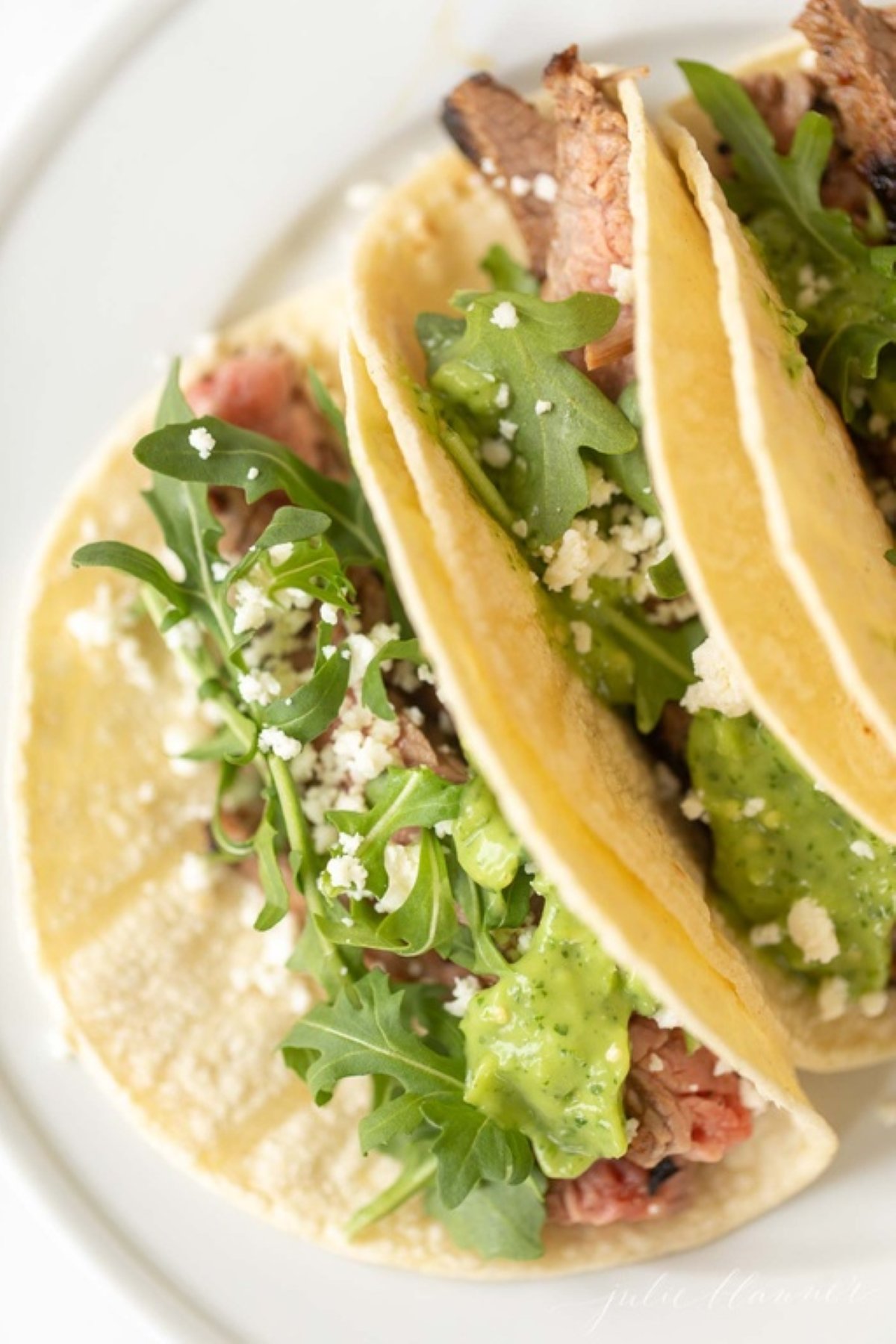 A white plate featuring three carne asada tacos topped with sauce, cheese and arugula.