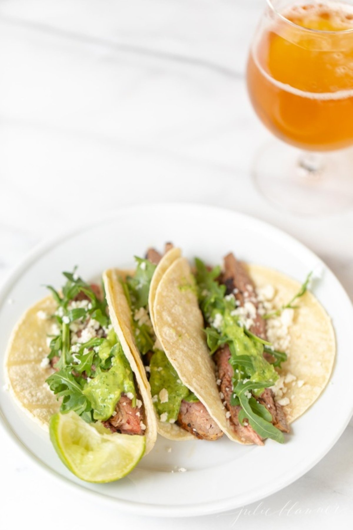 A white plate featuring three carne asada tacos topped with sauce, cheese and arugula.