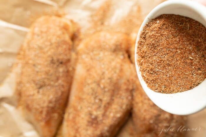 Three raw chicken breasts covered in blackened seasoning, bowl of spices in foreground.