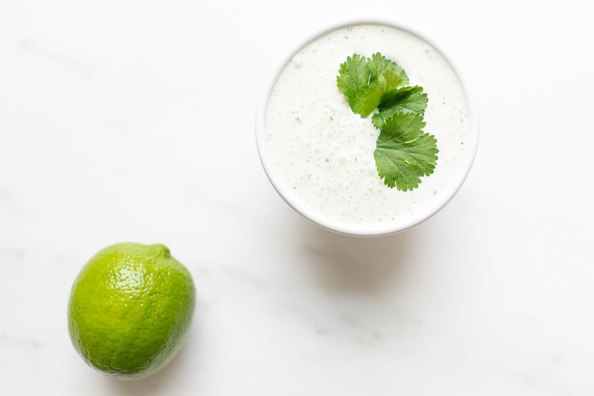 A white bowl of crema de cilantro on a white countertop, garnished with cilantro, lime to the side.