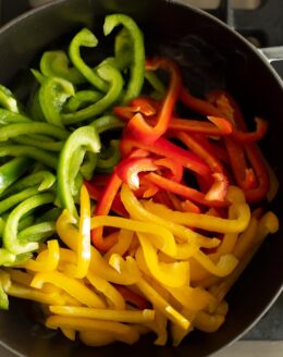 A cast iron pan on the stove, full of red green and yellow sliced peppers preparing to saute.