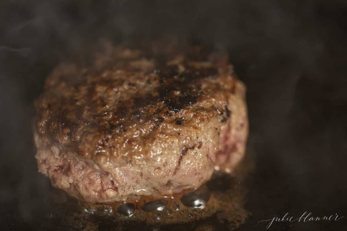 A cast iron pan with a burger patty cooking.