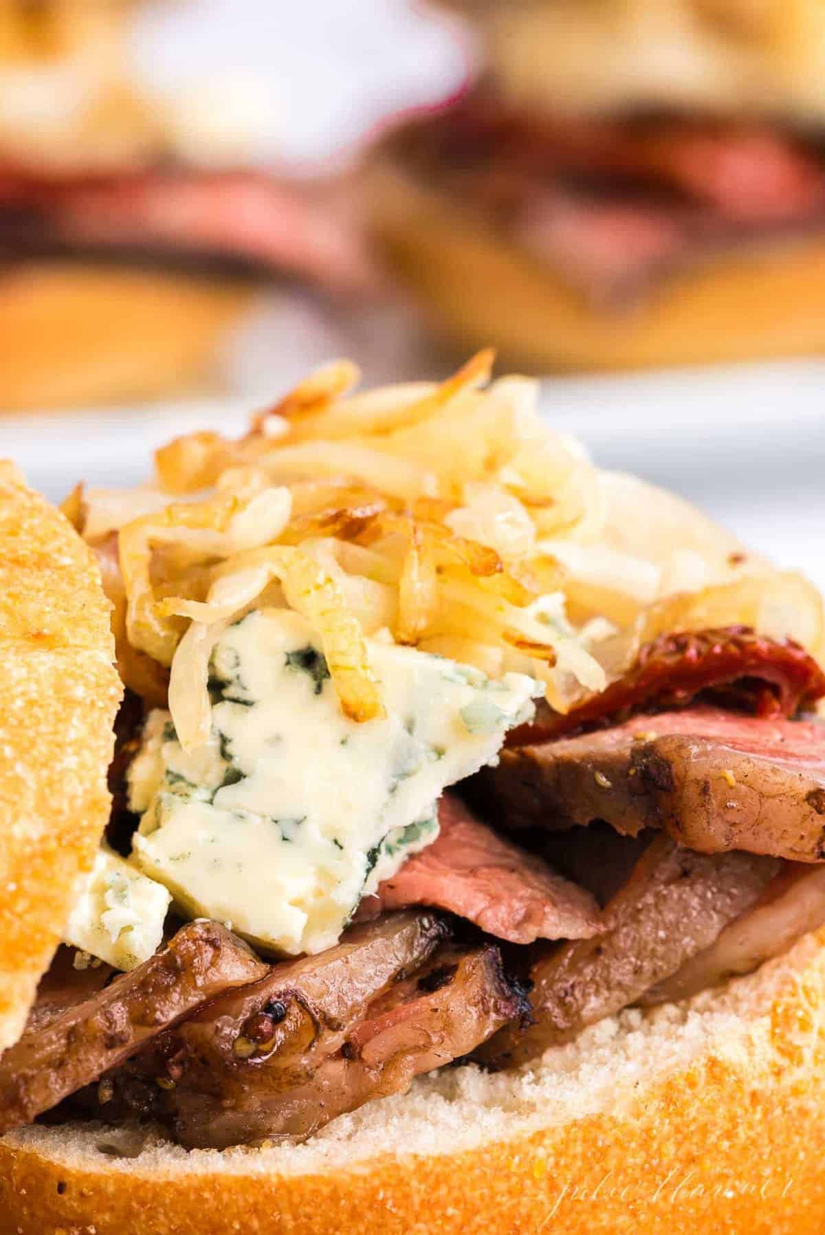 A steak sandwich with onion straws and cheese on top, top bun balanced to the side.