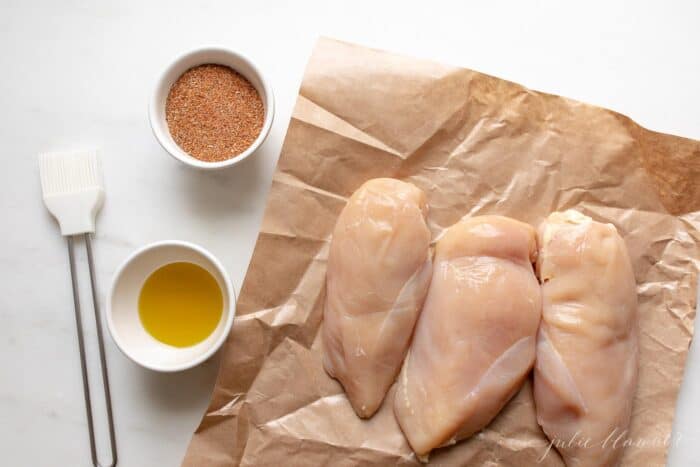 Butcher paper with three raw chicken breasts, a bowl of butter and blackened seasoning to the left.