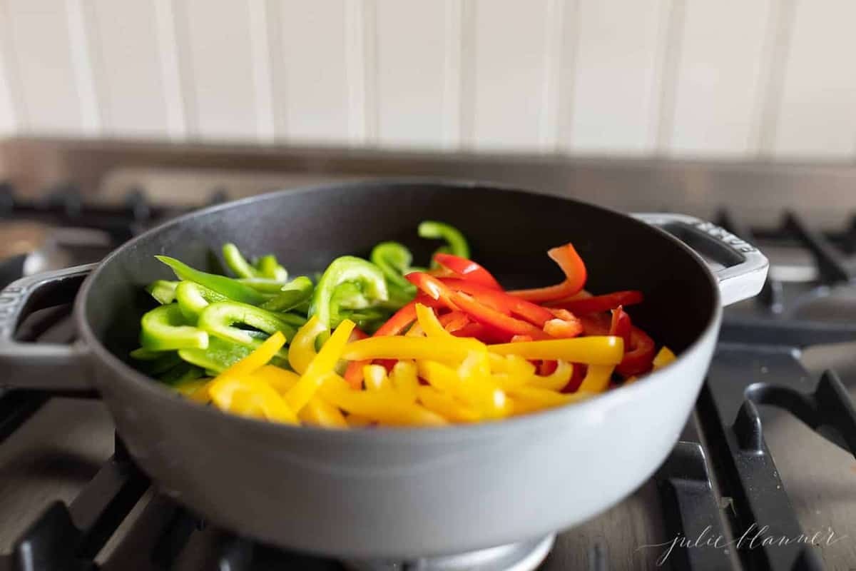 A cast iron pan on the stove, full of red green and yellow sliced peppers preparing to saute.