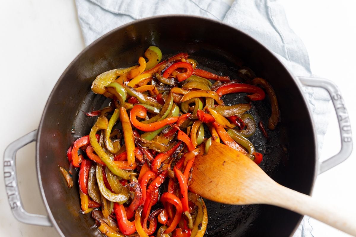 A cast iron pan full of sauteed bell peppers, wooden spoon to the side.