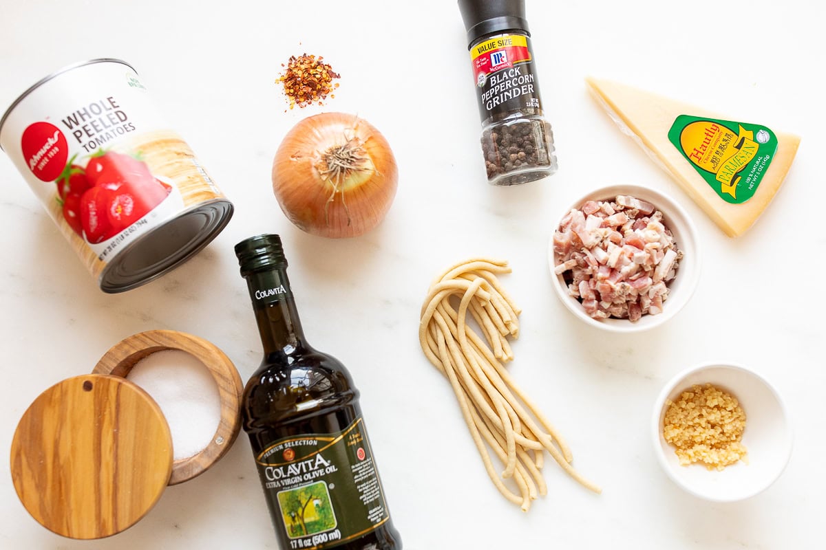 Ingredients for amatriciana sauce arranged on a white surface, including canned tomatoes, onion, black pepper, cheese, olive oil, diced bacon, and garlic.