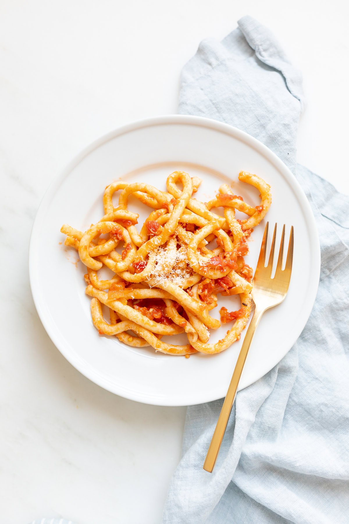 A white plate of bucatini all'amatriciana, alongside a gold fork on a light blue napkin.