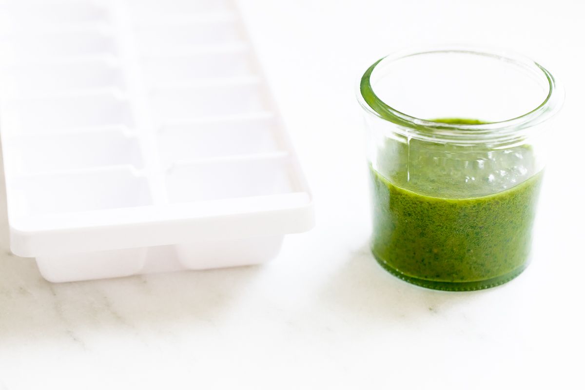 A small glass of basil pesto on a white marble countertop, with a white ice cub tray next to it.