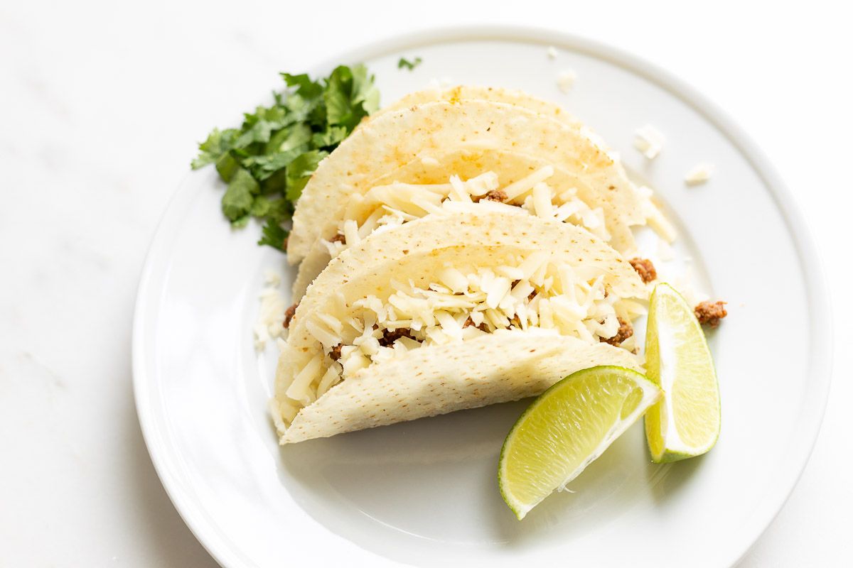 Beef tacos on a white plate, garnished with cheese, sour cream, and limes and cilantro on the plate.