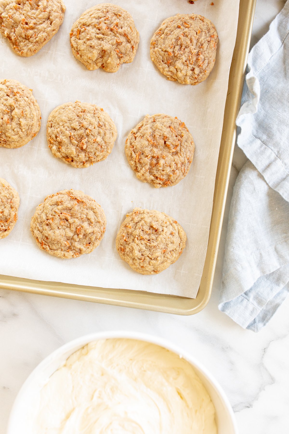 Carrot cake cookies on a gold baking sheet, bowl of frosting in the background.