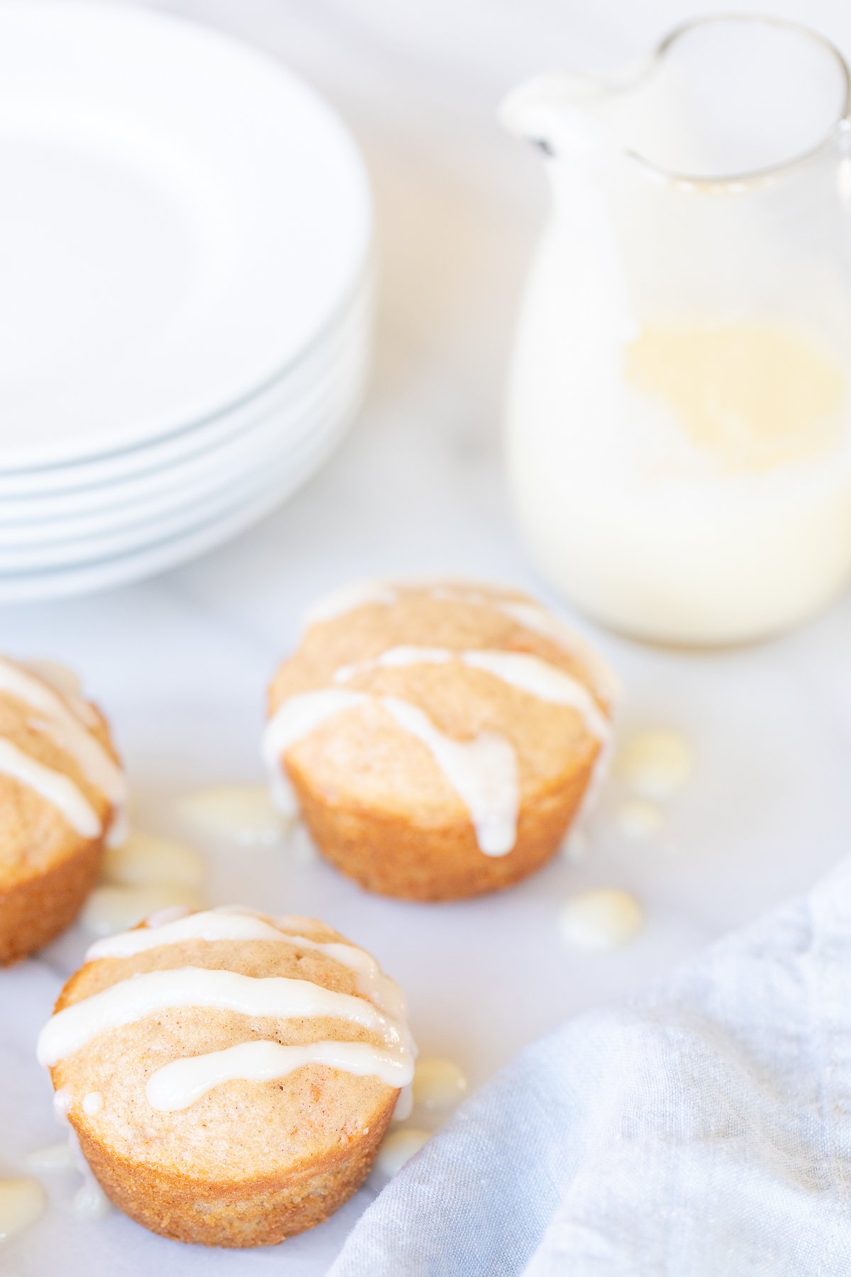 carrot cake muffins with a stack of plates and pitcher of frosting in the background.