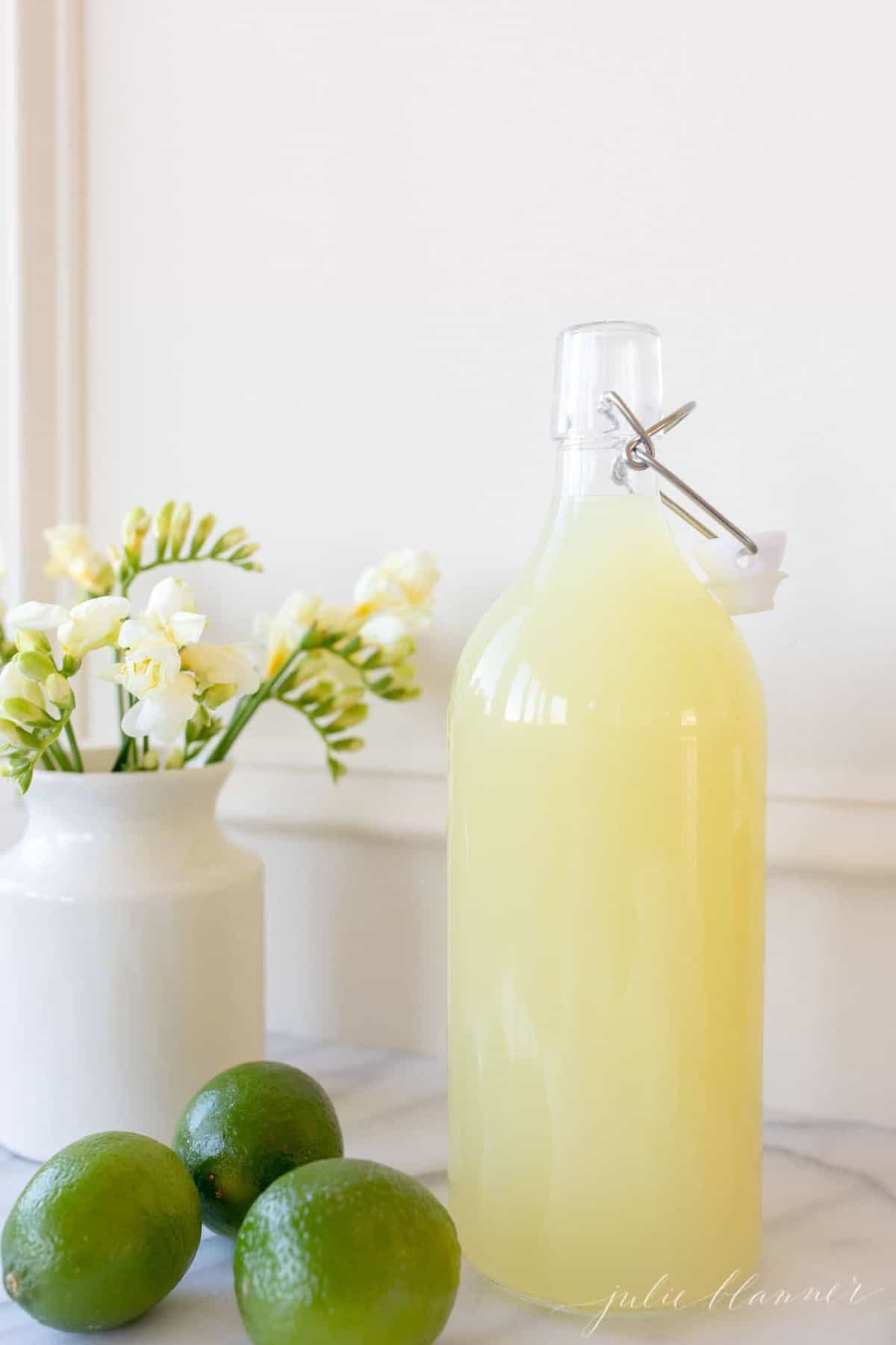 Homemade margarita mix in a glass carafe; a vase full of yellow flowers sits to the side. 