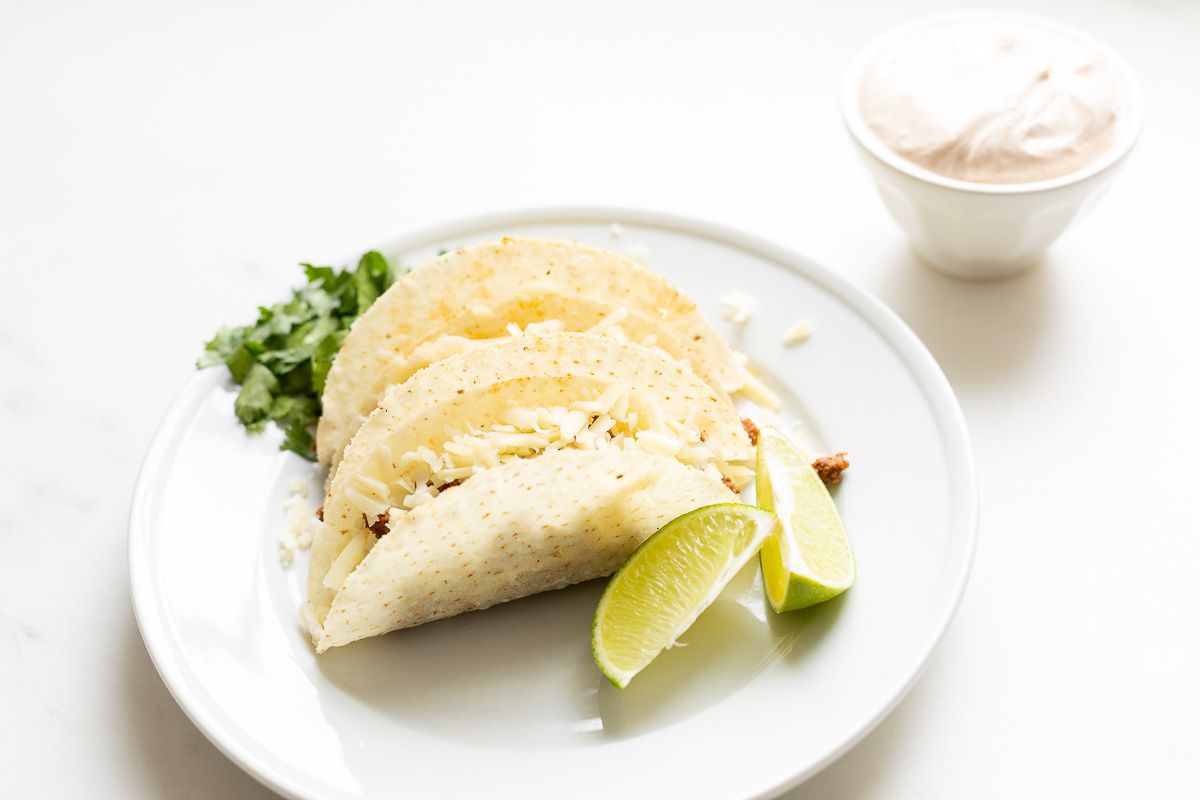 Beef tacos on a white plate, garnished with cheese, sour cream, and limes and cilantro on the plate.