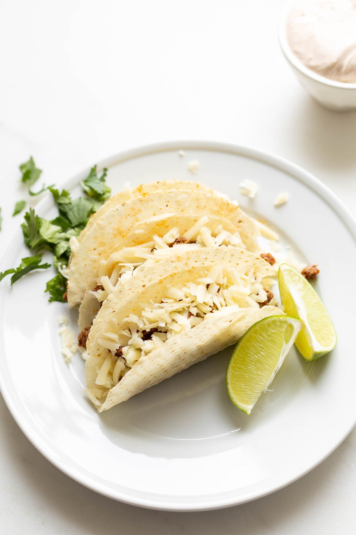 Beef tacos on a white plate, garnished with cheese, sour cream, and limes and cilantro on the plate.